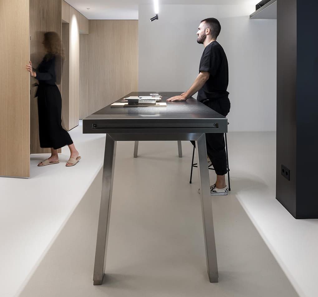 A man stands at a sleek, modern table in a minimalist room, while a woman walks by wooden panels in the background.
