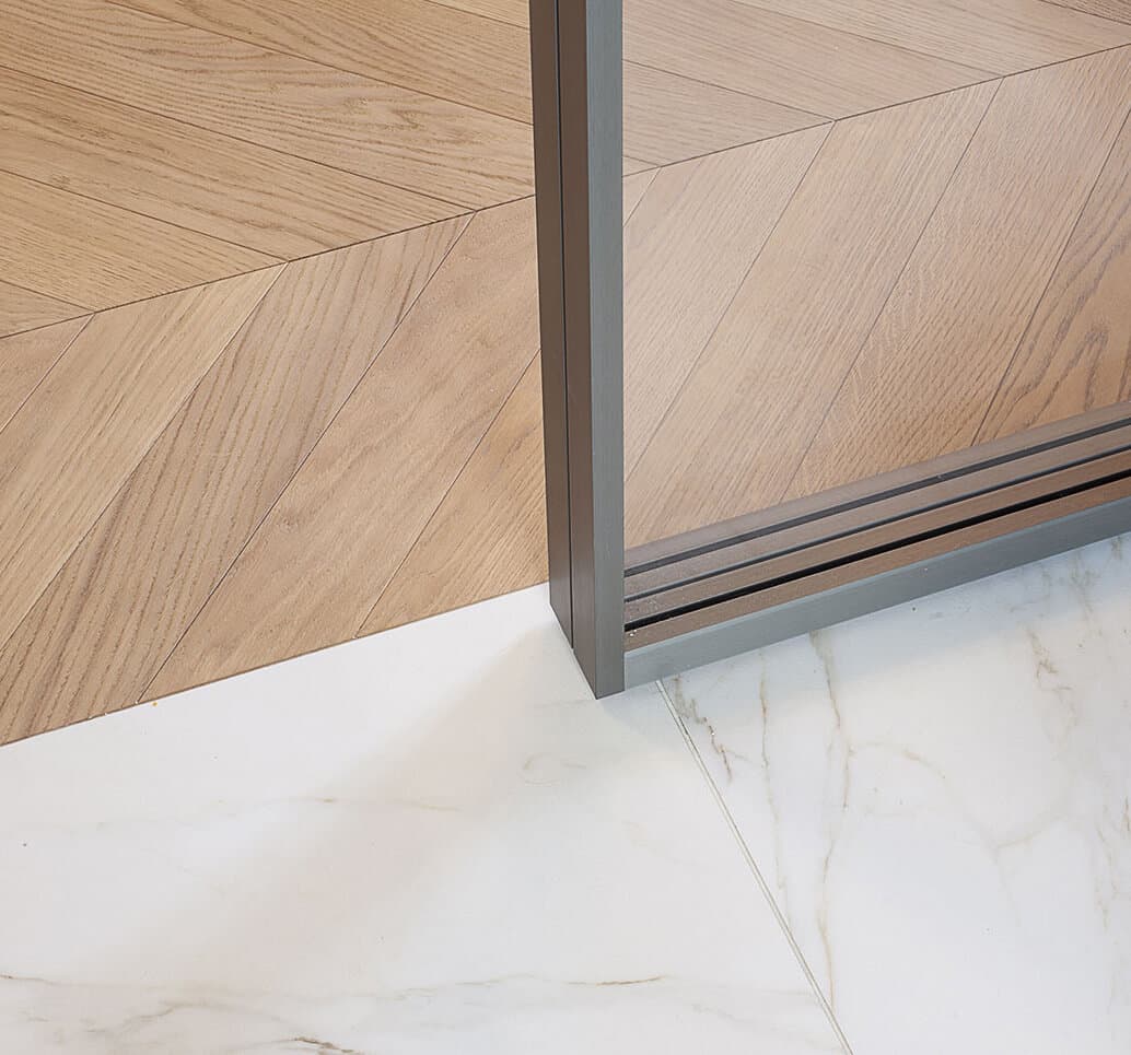 Close-up of a modern floor transition between light wood herringbone and white marble tiles, divided by a sleek metal frame.