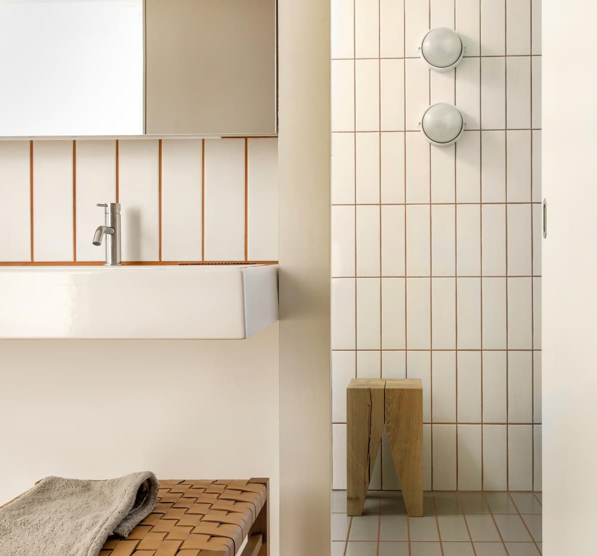Minimalist bathroom with white tiles, wooden stool, woven bench, and wall-mounted sink. Two round lights adorn the shower wall.