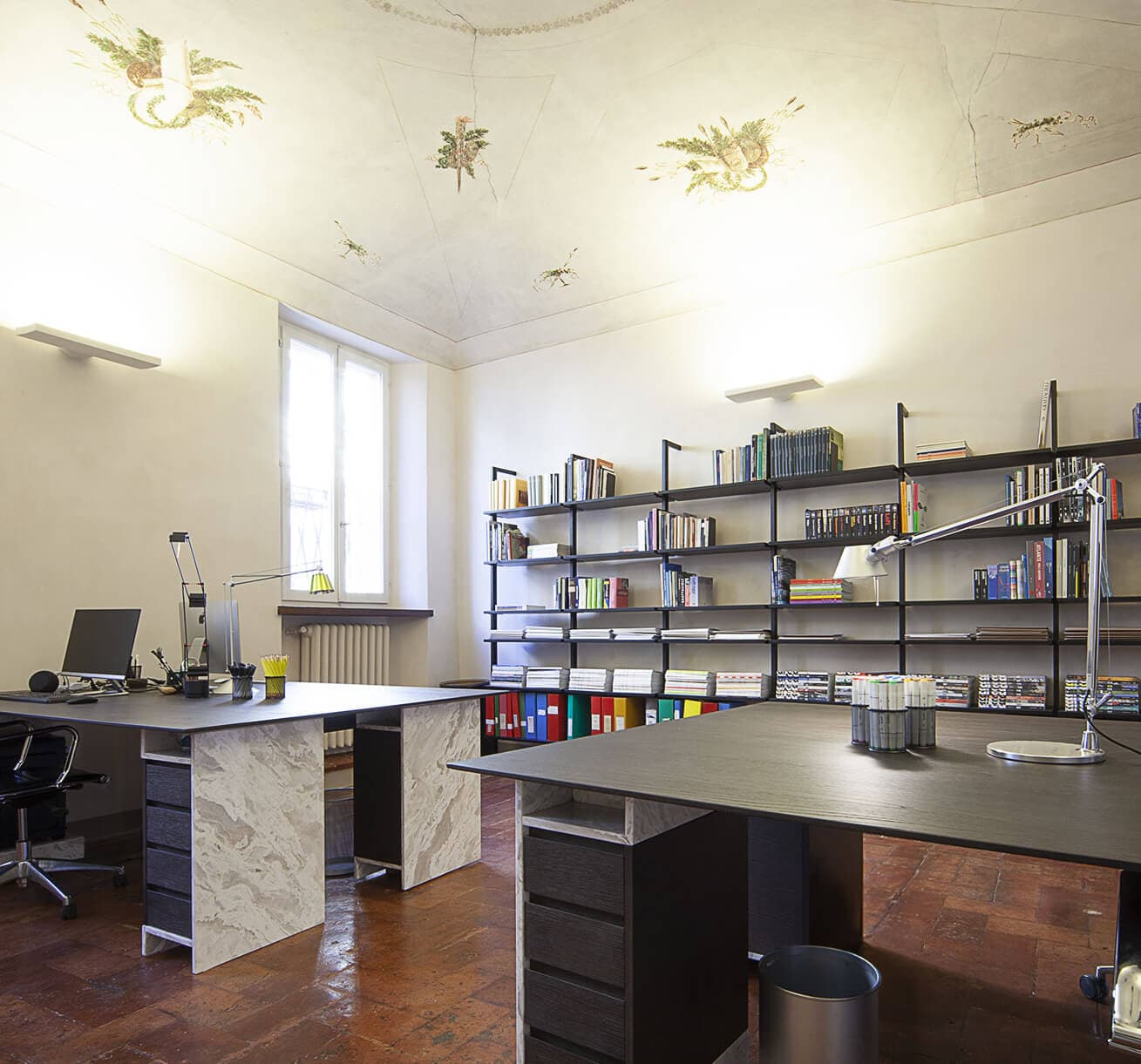 Modern office with two marble desks, shelves filled with books, decorative ceiling, and a large window letting in natural light.