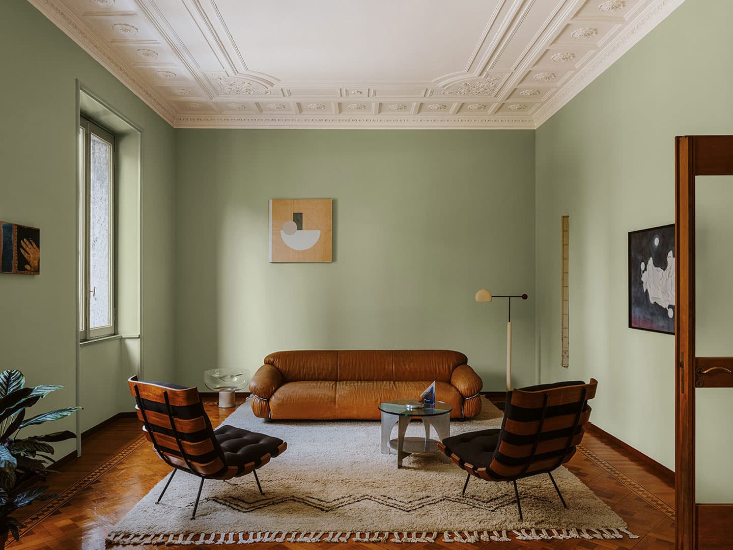 A minimalist living room with green walls, a brown sofa, two wooden chairs.