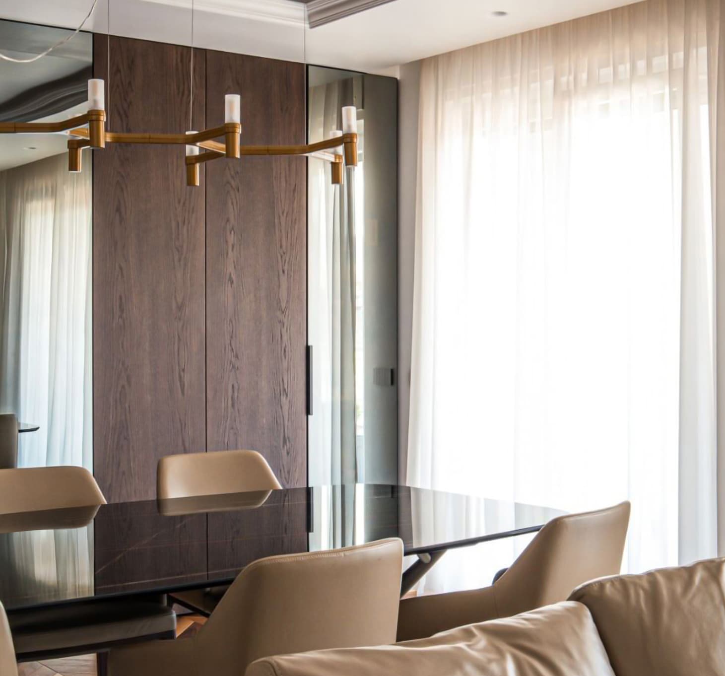 Modern dining room with a sleek black table, beige chairs, and a gold chandelier. Floor-to-ceiling windows with sheer curtains allow natural light.
