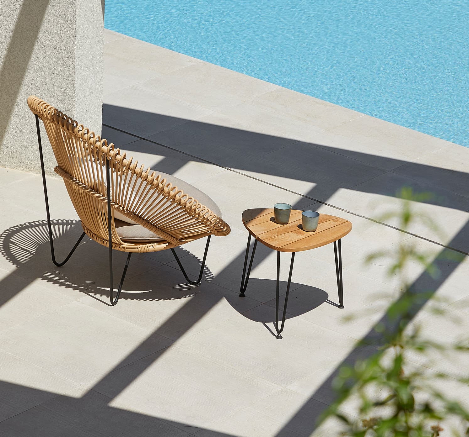 A wicker chair and small wooden table with two cups sit on a sunlit patio beside a clear blue pool, casting shadows on the tiled floor.