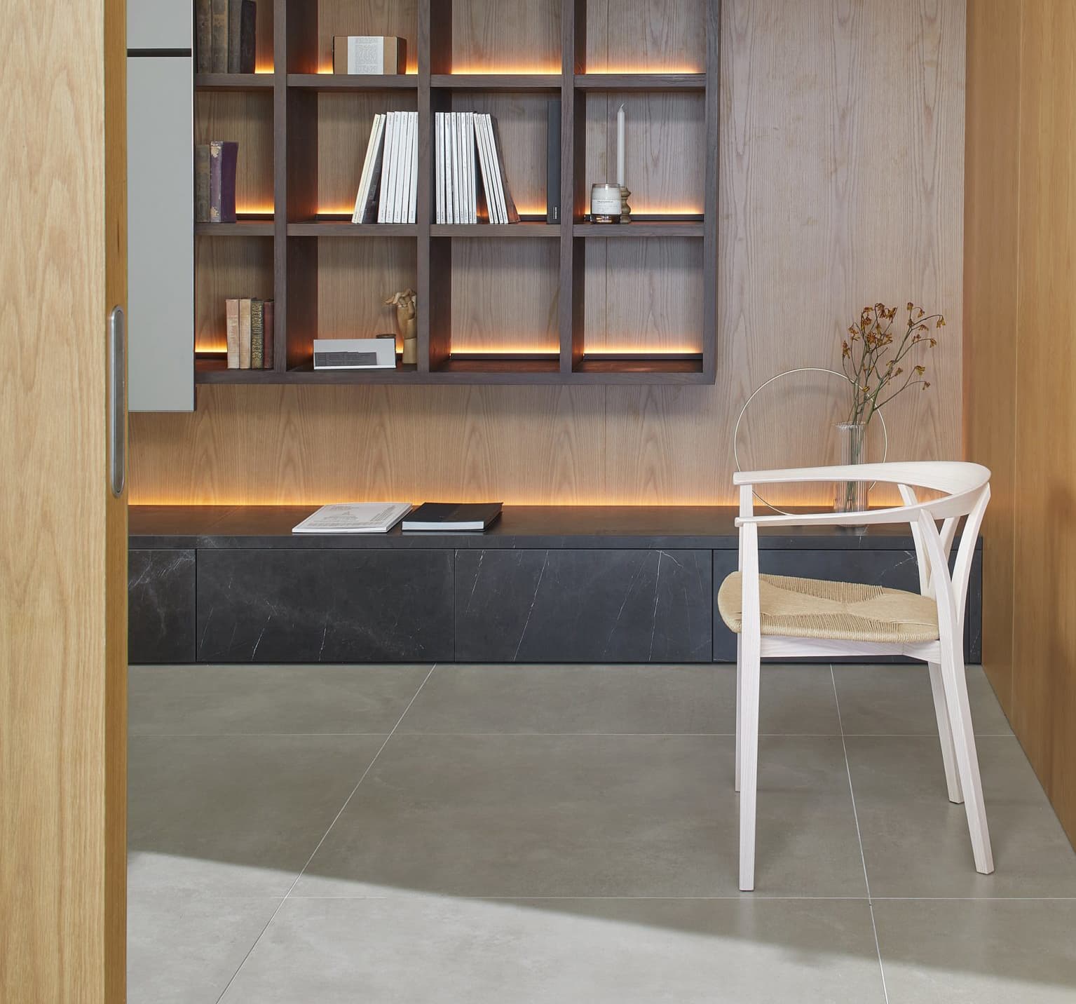 Minimalist room with a wooden bookshelf, illuminated shelves, a white chair, and a black bench. Neutral tones and simple decor create a calm atmosphere.