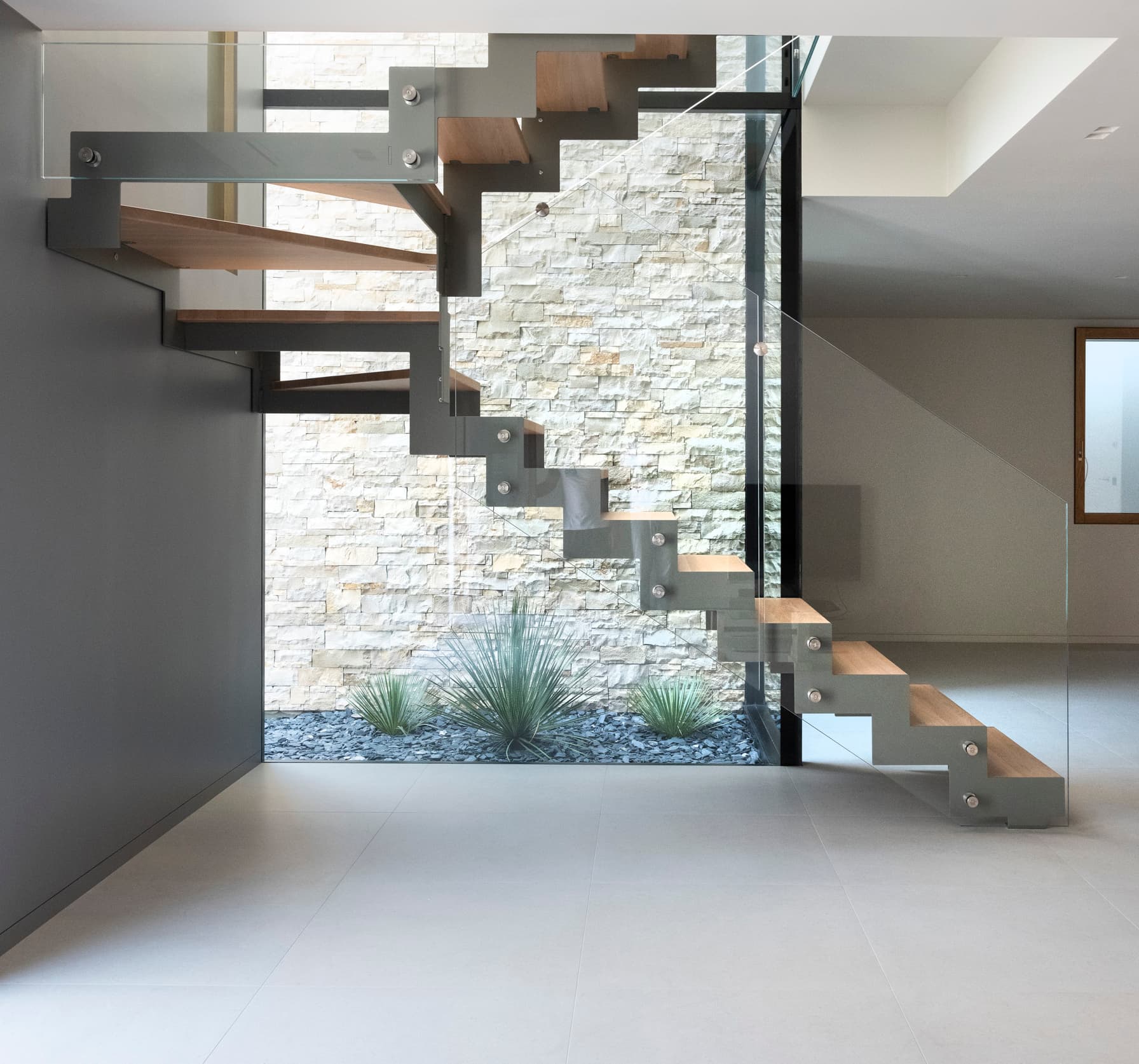 Modern interior with a floating wooden staircase, metal supports, and glass railing, set against a stone wall with plants below.