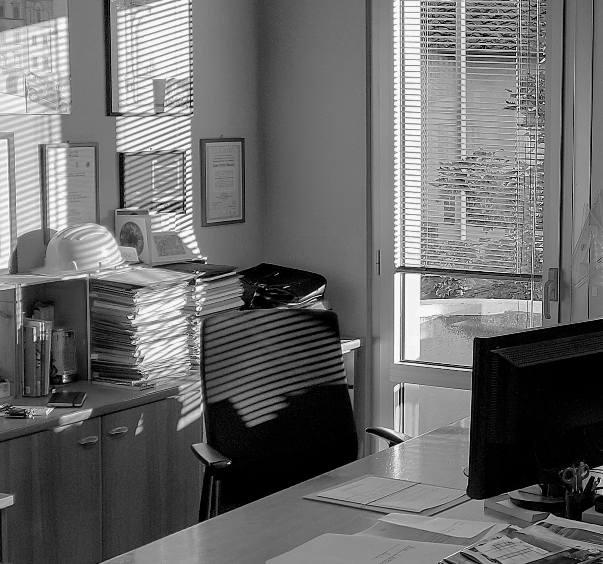 Sunlit office with a desk, chair, and computer. Blinds cast striped shadows on walls and furniture. Stacks of papers and hard hats on shelves.