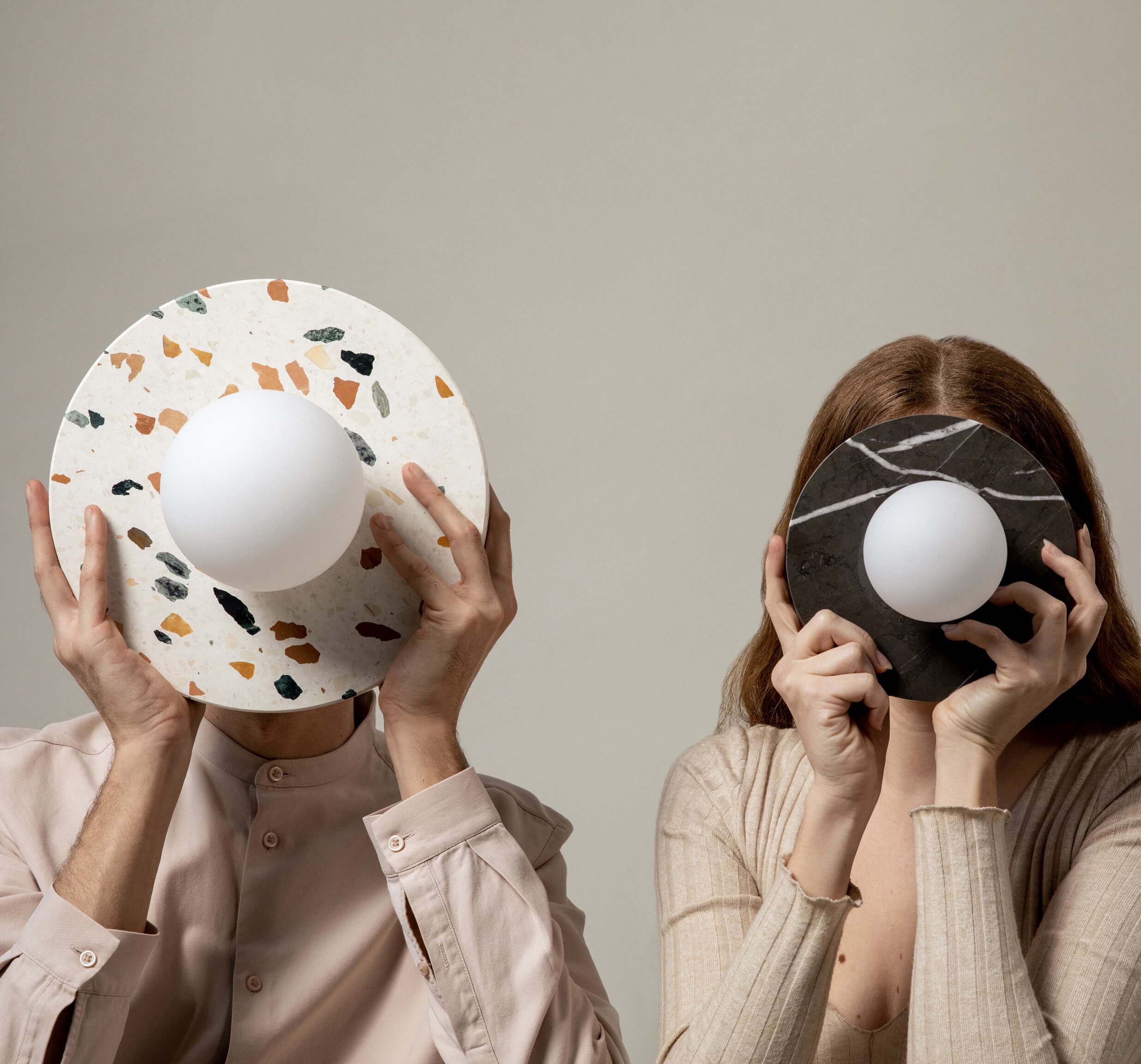Two people hold round, textured objects with central white spheres in front of their faces, set against a plain background.