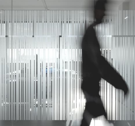 Silhouetted person walking past a frosted glass wall with vertical lines, creating a blurred effect in a modern interior setting.