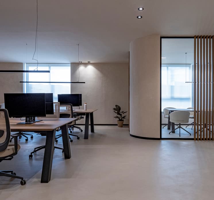 Modern office with minimalist design, featuring desks with computers, swivel chairs, and a glass-walled meeting room in the background.