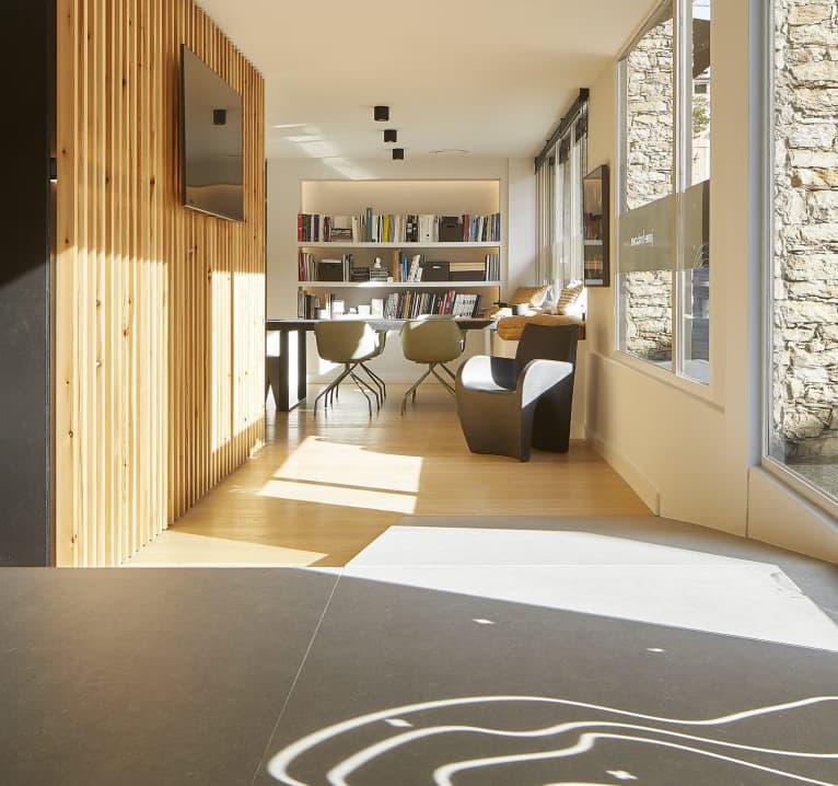 Modern room with wooden slat wall, large windows, bookshelves, and seating area. Sunlight casts shadows on the floor.