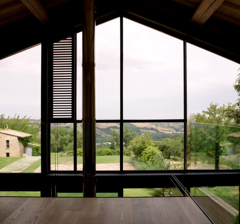 Large window with a view of a rural landscape, featuring rolling hills, a house, and lush greenery under a cloudy sky.