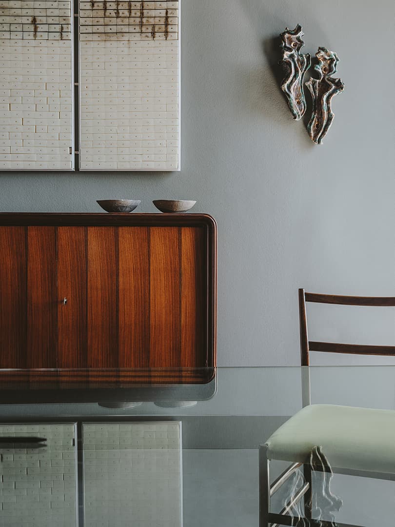Minimalist interior with a wooden cabinet, glass table, modern wall art, and a chair with a light green cushion against a gray wall.